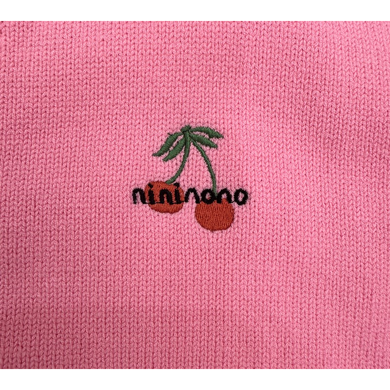 Detailed shot of the playful cherry embroidery patch stitched onto the chest of the pink cardigan.