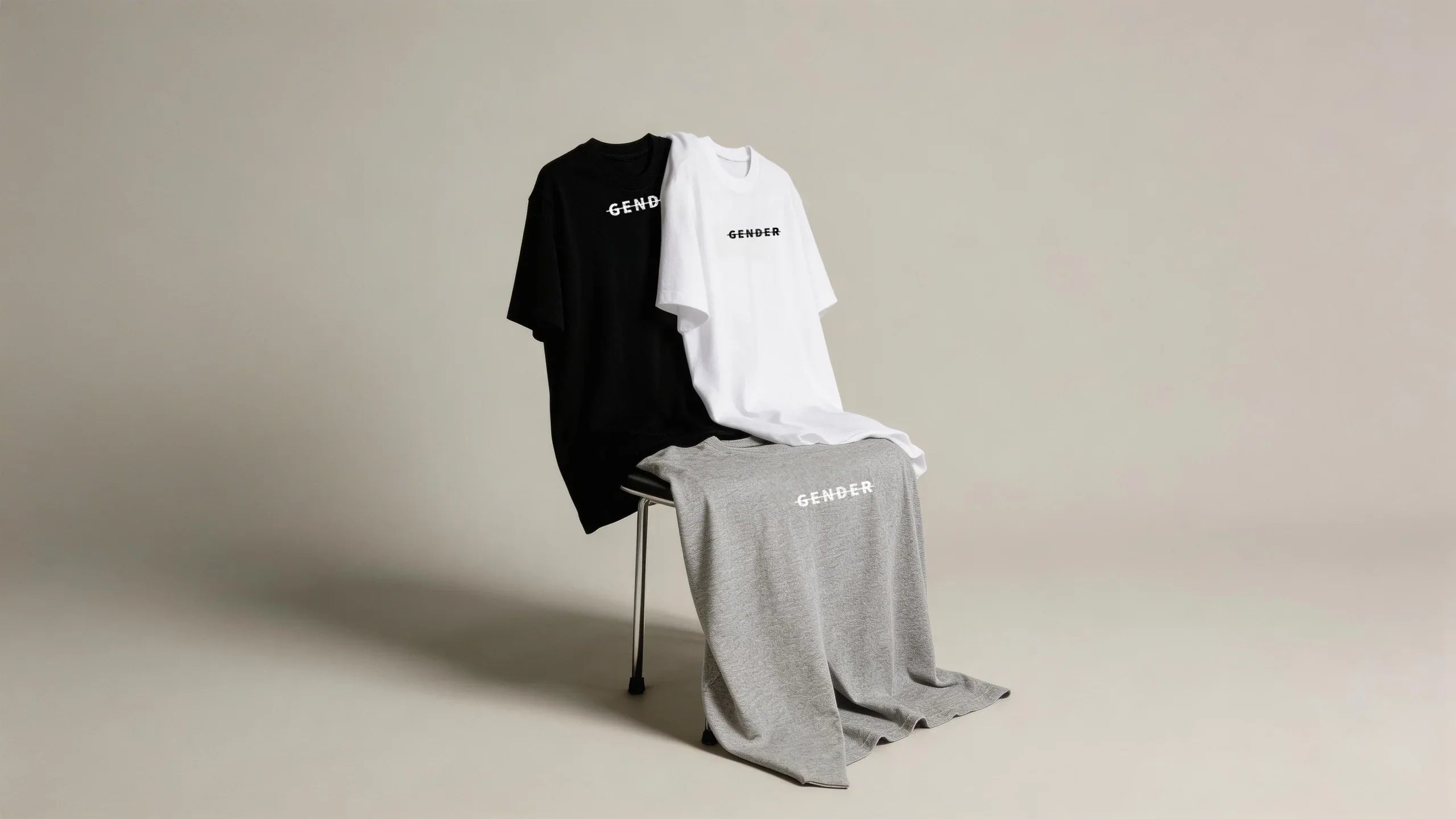Three OGENDER standard-fit 230g cotton t-shirts in black, white, and grey displayed on a chair, featuring the gender-neutral logo.