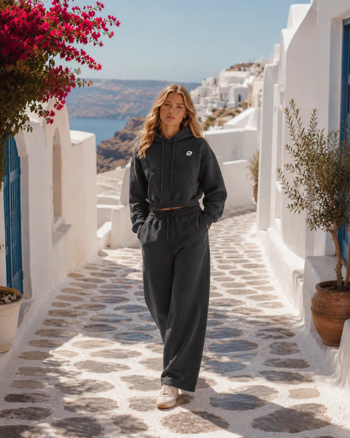 Russian female model wearing a cropped dark grey hoodie and wide-leg sweatpants walking through a sunlit Santorini alley.