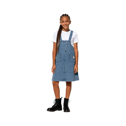 A young girl modeling the indigo denim pinafore dress, showing the waist pocket placement.