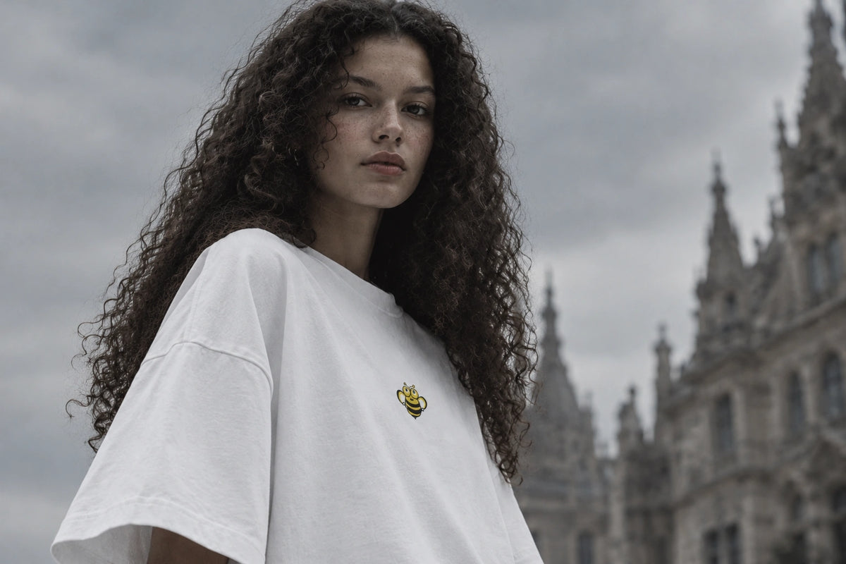 OGender Insect Collection oversized white T-shirt featuring a small yellow cartoon bee print, worn by a curly-haired female model against a cool-toned blurred architectural background.