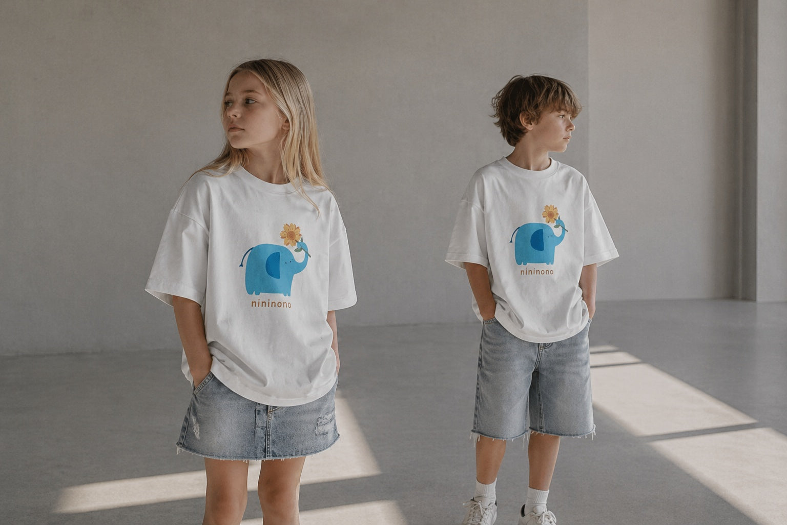 Two kids wearing ogender 100% cotton 230g heavyweight oversized white t-shirts featuring a cute blue elephant and sunflower graphic, paired with denim, in a bright minimalist studio.