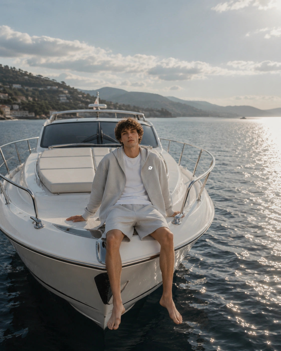 Curly-haired male model wearing a light grey Ogender zip hoodie with a white T-shirt and matching sweat shorts, sitting barefoot on the bow of a yacht under the summer sun in a relaxed coastal setting.