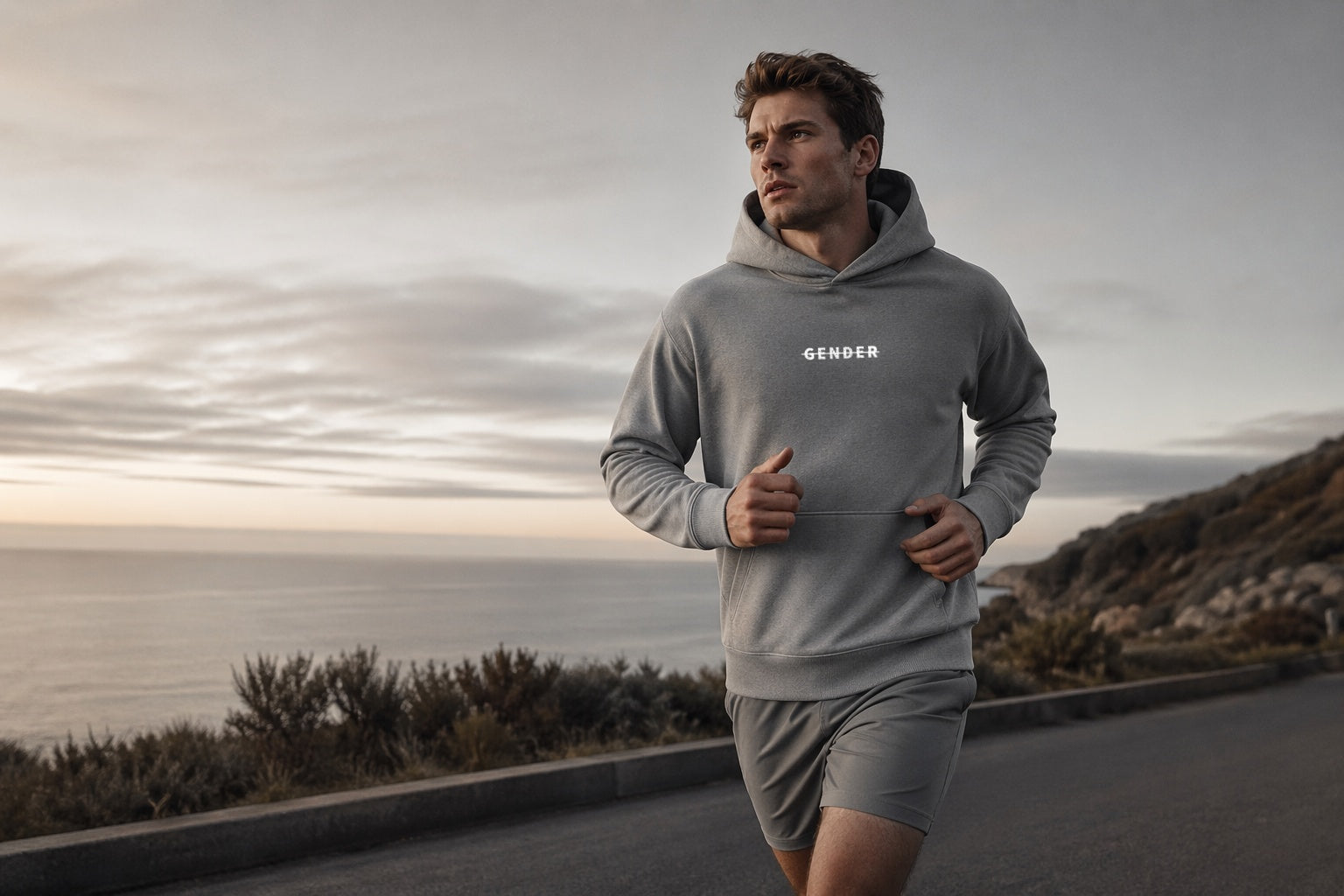 A male model jogging on a quiet coastal road during sunrise, wearing an Ogender grey drawstring-free hoodie and matching running shorts, representing a clean and healthy lifestyle.