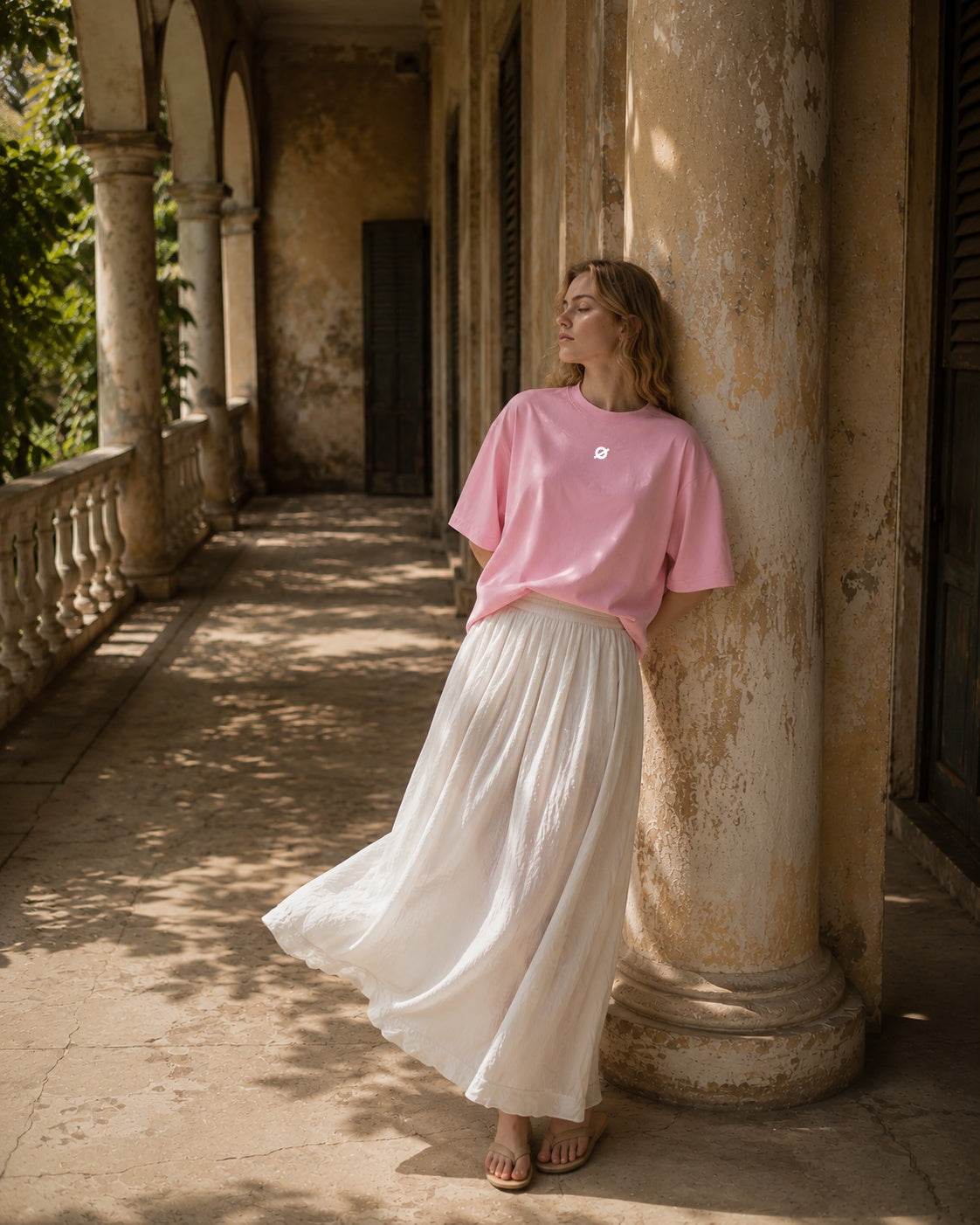 A model wearing an ogender pink oversized t-shirt and a flowing white maxi skirt, leaning against a vintage stone pillar in a sunlit historic balcony.