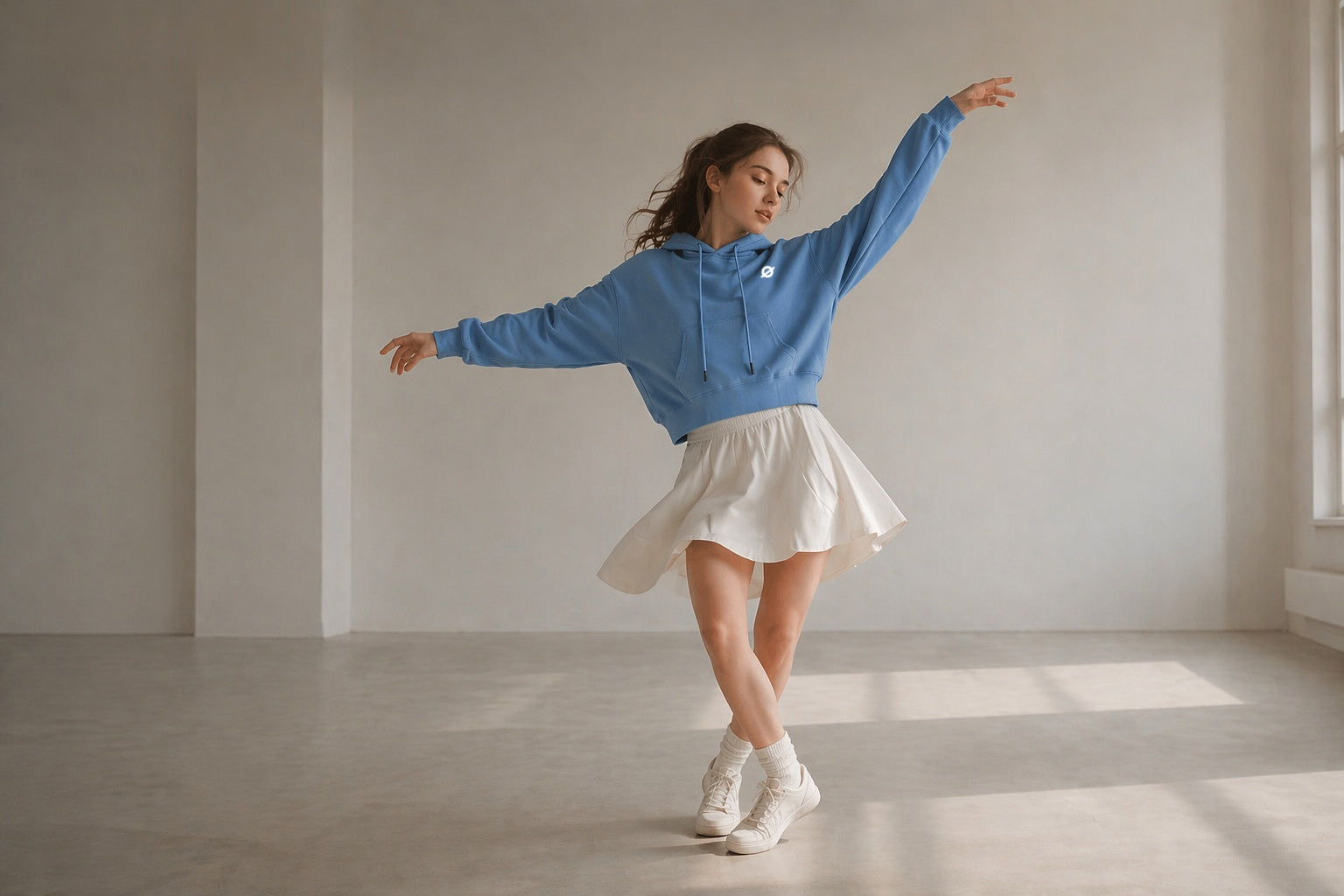 A female dancer posing in an Ogender sky blue cropped drawstring hoodie and a white flared skirt in a minimalist sunlit studio, showcasing the high-waist fit and lightweight fabric.