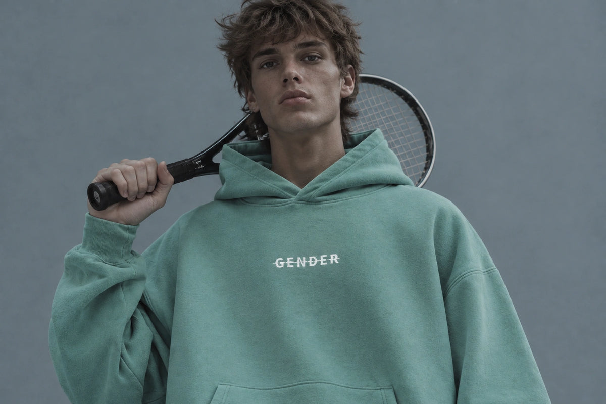 OGender oversized teal fleece pullover hoodie with a white chest logo, worn by a young male model holding a tennis racket against a cool gray-blue studio background.